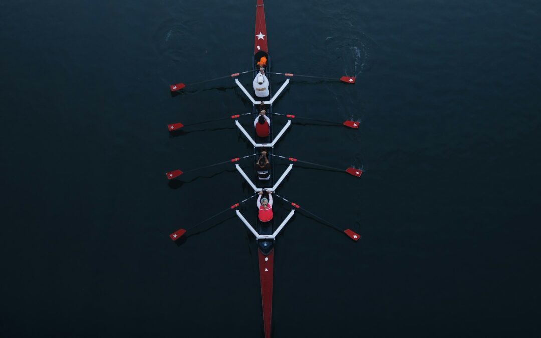 A four-person rowing crew seen from directly above on dark water, oars in unison